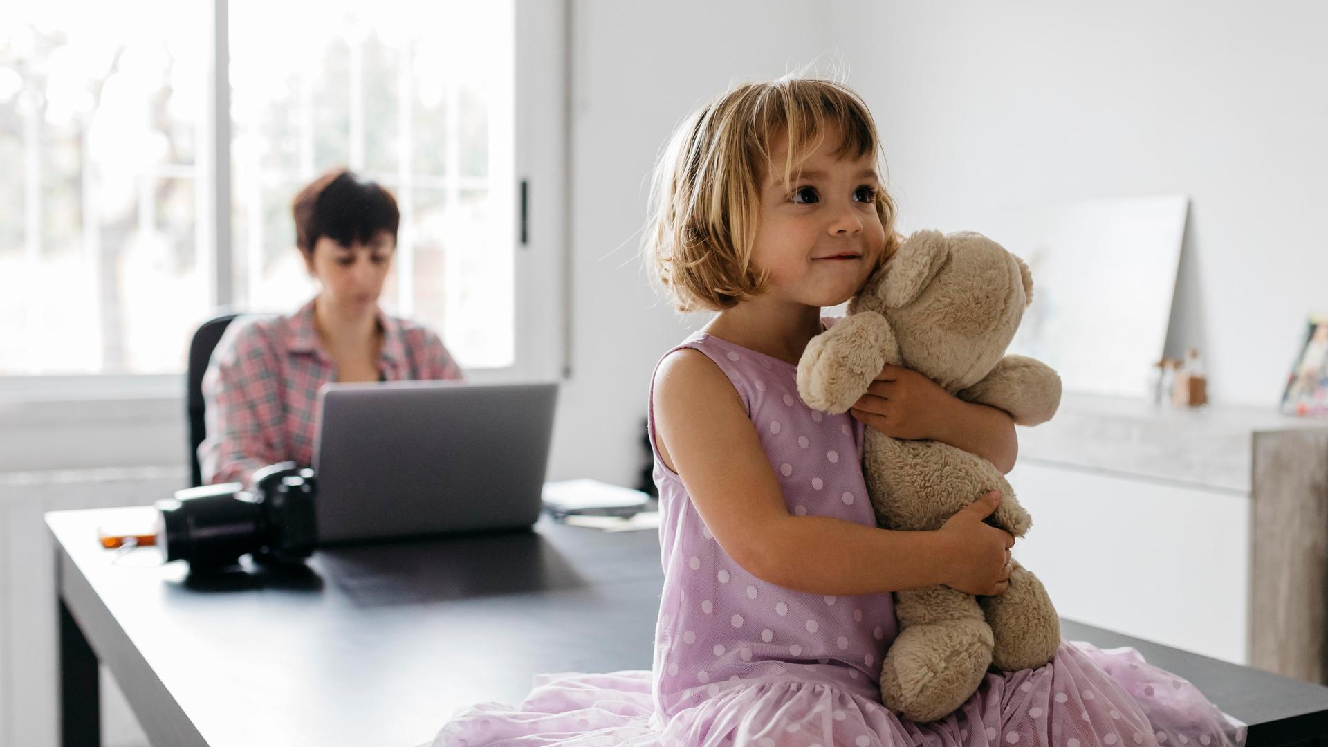 Eine Frau arbeitet an einem Tisch mit dem Laptop. Auf dem Tisch sitzt ein Mädchen, das einen Teddybären im Arm hält. Eine Frau arbeitet an einem Tisch mit dem Laptop. Auf dem Tisch sitzt ein Mädchen, das einen Teddybären im Arm hält.