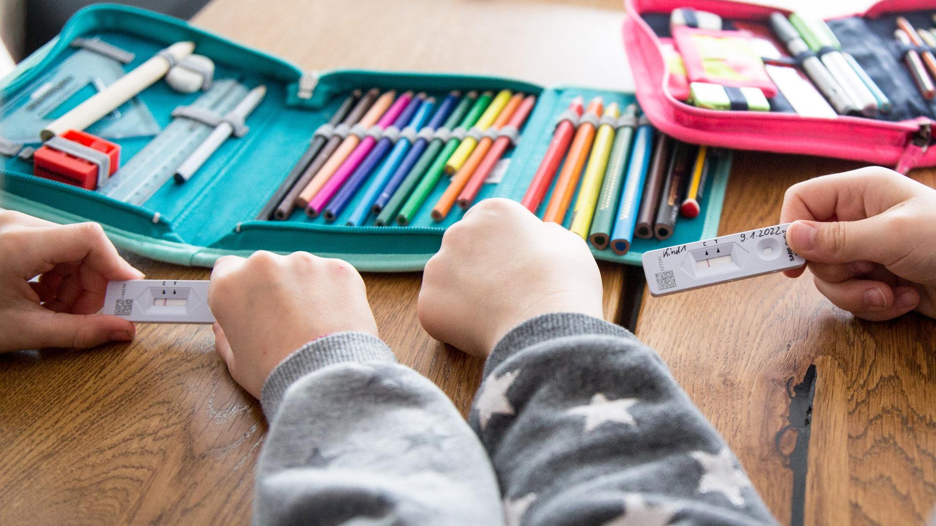 Zwei Schulkinder halten ihren SARS-CoV-2 Rapid Antigen Test in der Hand dieser Negativ anzeigt, im Hintergrund liegt eine Federmappe, Hefte und Bücher für die Schule auf dem Tisch. Zwei Schulkinder halten ihren SARS-CoV-2 Rapid Antigen Test in der Hand dieser Negativ anzeigt, im Hintergrund liegt eine Federmappe, Hefte und Bücher für die Schule auf dem Tisch.