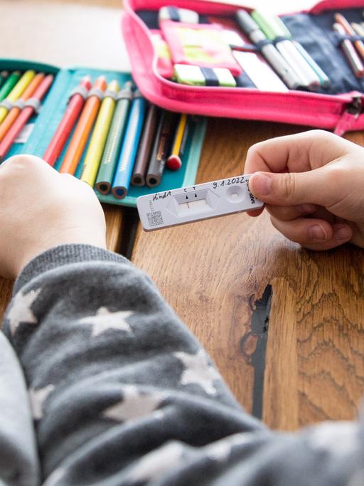 Zwei Schulkinder halten ihren SARS-CoV-2 Rapid Antigen Test in der Hand dieser Negativ anzeigt, im Hintergrund liegt eine Federmappe, Hefte und Bücher für die Schule auf dem Tisch.