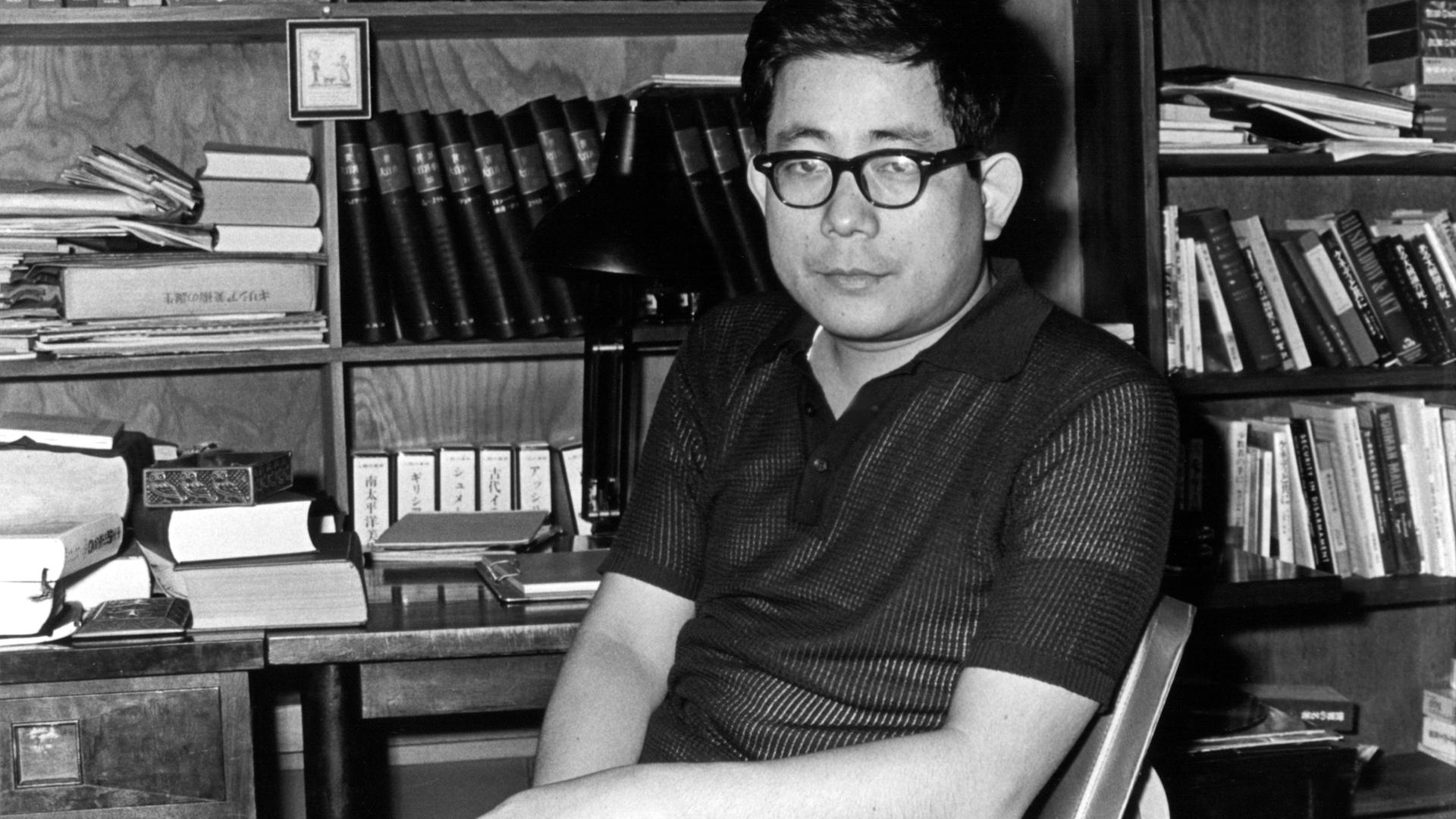 Schwarzweißfoto von Kenzaburo Oe, der als junger Mann an seinem Schreibtisch sitzt. Im Hintergrund ein Bücherregal. Schwarzweißfoto von Kenzaburo Oe, der als junger Mann an seinem Schreibtisch sitzt. Im Hintergrund ein Bücherregal.