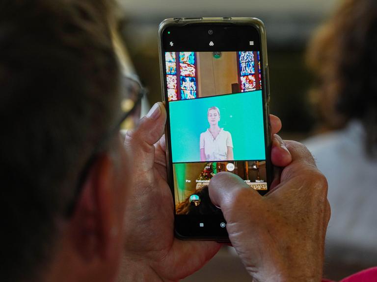 Ein Mann hält ein Smartphone in der Hand, auf dem eine KI-generierte Predigt abgespielt wird.