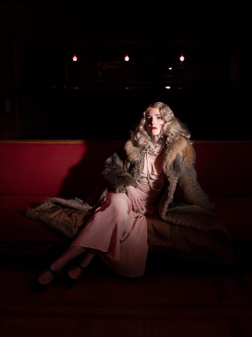 Szenenfoto aus dem Stück "Hyäne Fischer – Das totale Musical" der Berliner Volksbühne. Zu sehen ist eine elegante Frau mit blondem welligem Haar und Pelzmantel auf einem roten Sessel.