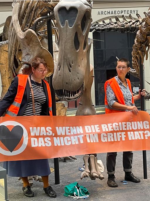 Zwei Klimaaktivistinnen mit einem Banner "Was, wenn die Regierung das nicht im Griff hat?" an den Haltestangen eines Dinosauriers festgeklebt. 
