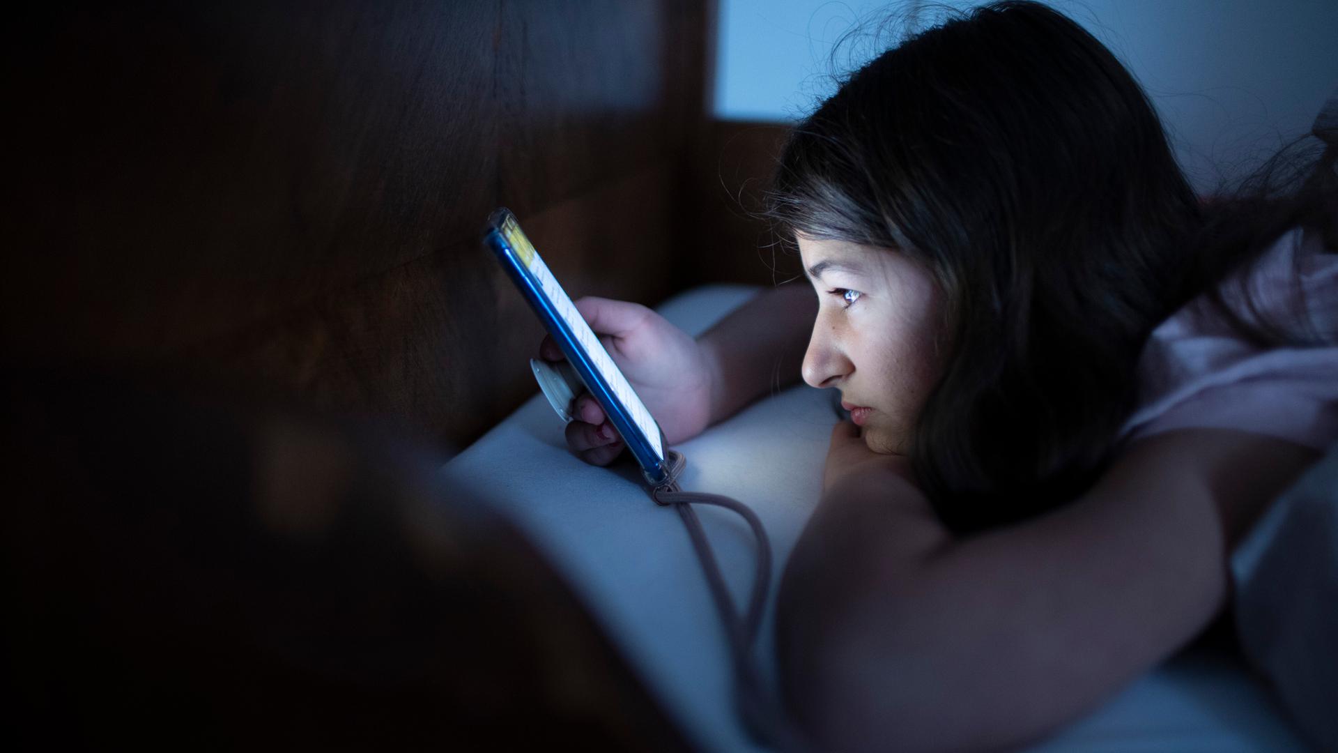 Ein Kind liegt mit einem Handy im Bett und schaut auf den Bildschirm. Ein Kind liegt mit einem Handy im Bett und schaut auf den Bildschirm.