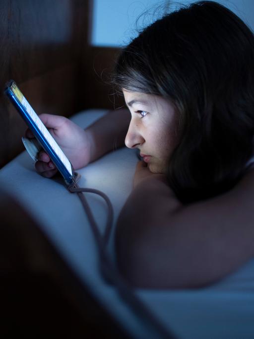Ein Kind liegt mit einem Handy im Bett und schaut auf den Bildschirm.