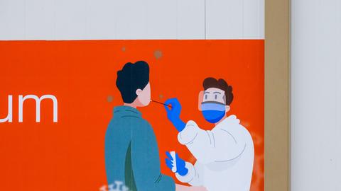 Auf einem Plakat vor einem Corona-Testzentrum ist zu sehen, wie ein Abstrich genommen wird.