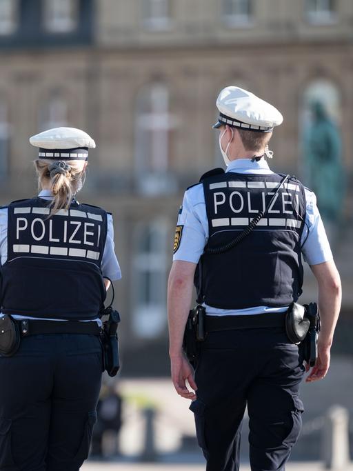Ein Polizist und eine Polizistin gehen über den Schlossplatz in Stuttgart, das Schloss ist im Hintergrund zu sehen. Sie haben dem Betrachter den Rücken zugewandt. 