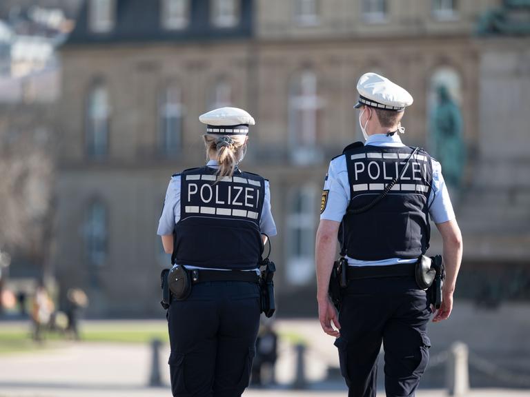Ein Polizist und eine Polizistin gehen über den Schlossplatz in Stuttgart, das Schloss ist im Hintergrund zu sehen. Sie haben dem Betrachter den Rücken zugewandt. 
