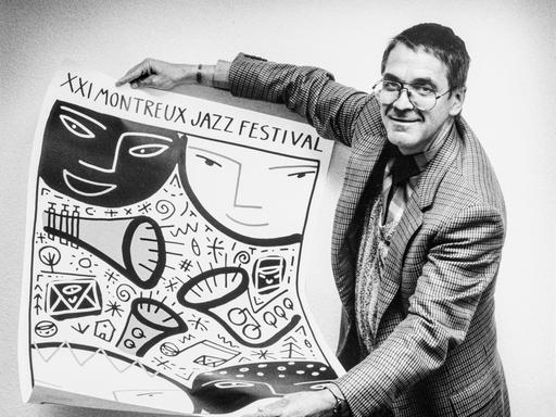 Claude Nobs hält auf dem Schwarz.Weiß-Bild das Plakat zum 21. Montreux Jazz Festival in den Händen und zeigt es in die Kamera. Er trägt das Haar kurz und hat eine Brille.