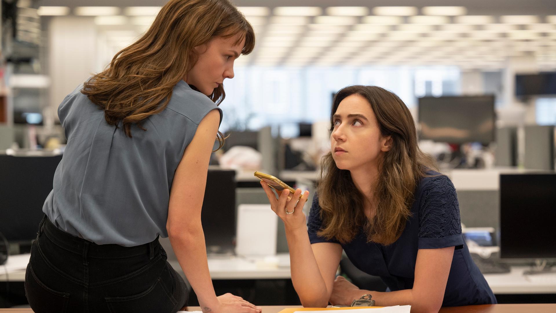Megan Twohey (Carey Mulligan) und Jodi Kantor (Zoe Kazan, v.l.) im Film She Said von Maria Schrader. Die Figur Megan Twohey sitzt auf dem Schreibtisch, Jodi Kantor lehnt daran. Sie hält ein Mobiltelefon. Beide schauen einander an. Im Hintergrund ist unscharf ein Großraumbüro zu sehen. Megan Twohey (Carey Mulligan) und Jodi Kantor (Zoe Kazan, v.l.) im Film She Said von Maria Schrader. Die Figur Megan Twohey sitzt auf dem Schreibtisch, Jodi Kantor lehnt daran. Sie hält ein Mobiltelefon. Beide schauen einander an. Im Hintergrund ist unscharf ein Großraumbüro zu sehen.