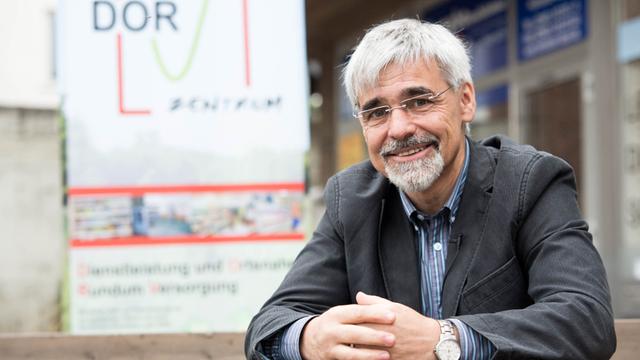Ein Mann mit grauen Haaren, Brille, Schnauzer und Kinnbart sitzt mit gefalteten Händen vor dem Schild eines dorfladens.
Ein Mann mit grauen Haaren, Brille, Schnauzer und Kinnbart sitzt mit gefalteten Händen vor dem Schild eines dorfladens.