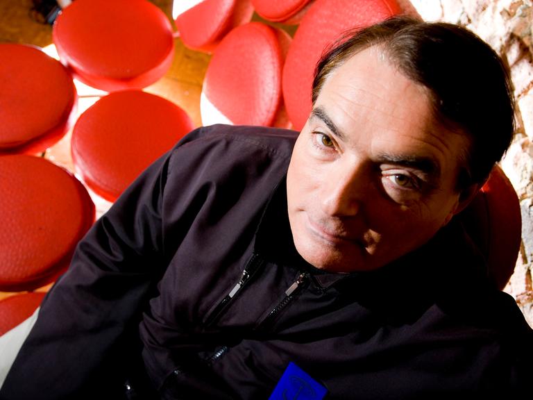 Porträt von Wolfgang Flür, dem Schlagzeuger von Kraftwerk. Er sitzt auf einem "Sofa", dass aus lauter roten Einzelsitzen besteht.