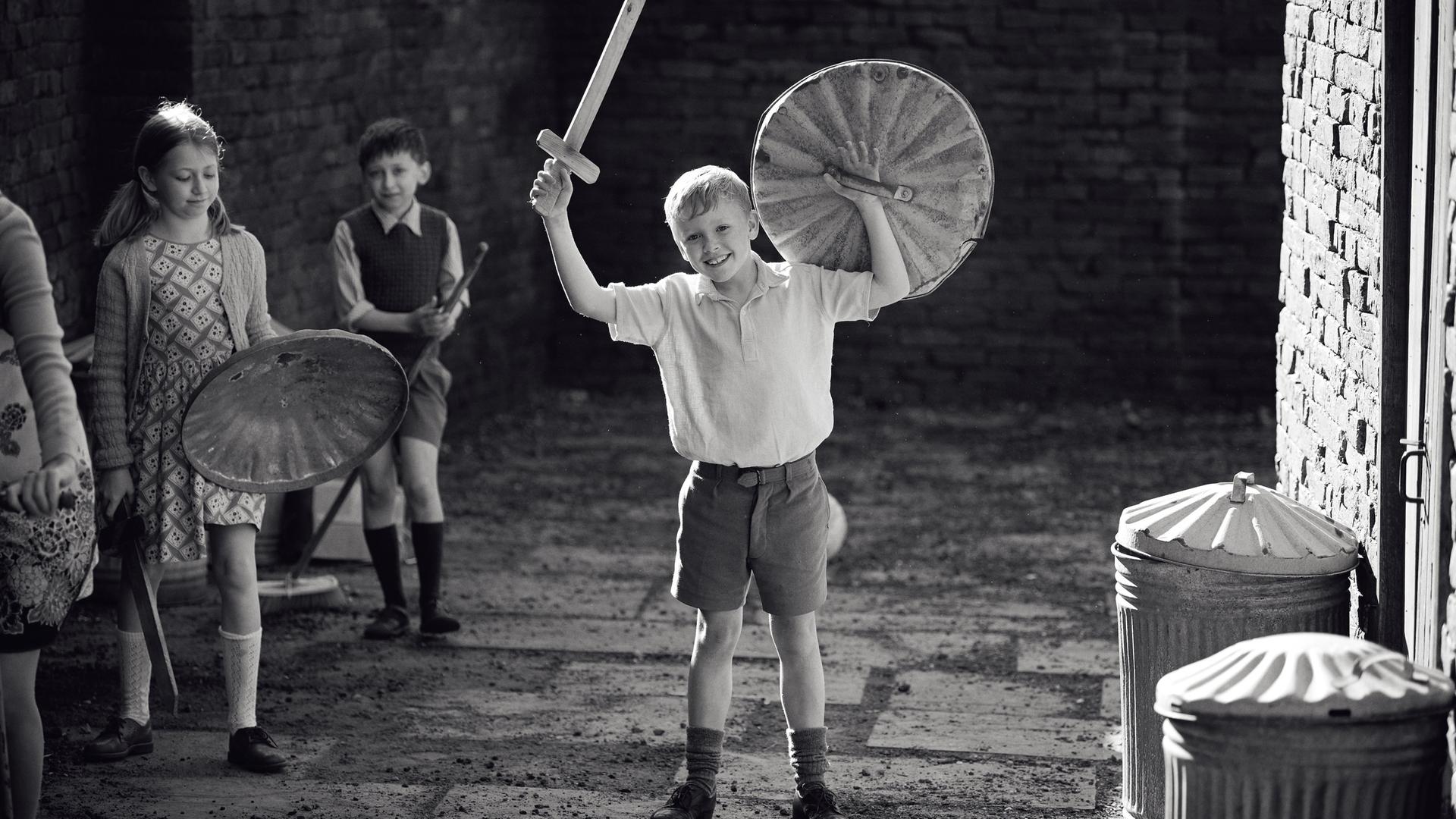 Im Still aus "Belfast" steht der kindliche Hauptdarsteller mit einem aus Holz gebastelten Schild und Schwert jubelnd auf einem Hof.