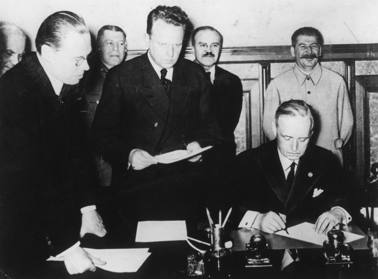 An einem Tisch sitzt Außenminister Joachim von Ribbentrop und unterzeichnet den Hitler-Stalin-Pakt. Am rechten Bildrand hinter ihm steht Josef Stalin.