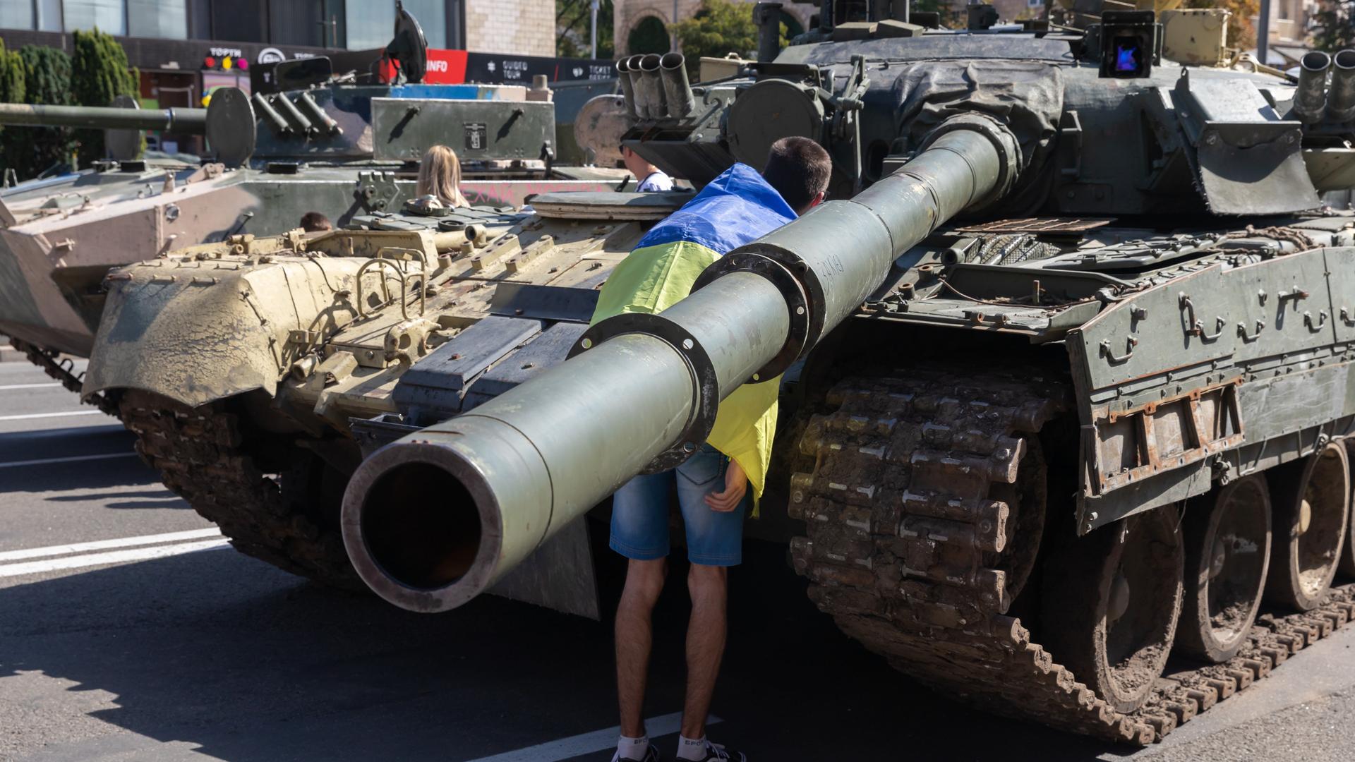 Ein in eine ukrainische Flagge gewickelter Mann vor einem zerstörten russischen Panzer 