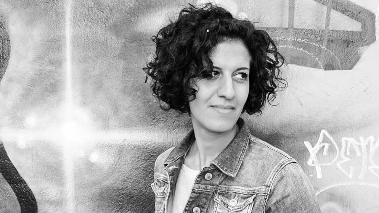 Die Schwarzweiß-Fotografie zeigt eine Frau mit lockigen, dunklen Haaren, die vor einer mit Graffiti besprühten Wand steht. Es die Musikerin und Kulturmanagerin Bahar Roshanai. Die Schwarzweiß-Fotografie zeigt eine Frau mit lockigen, dunklen Haaren, die vor einer mit Graffiti besprühten Wand steht. Es die Musikerin und Kulturmanagerin Bahar Roshanai.