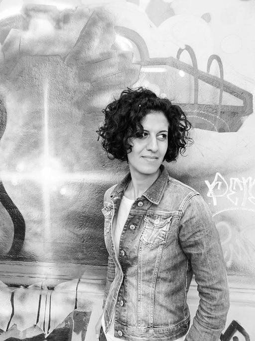 Die Schwarzweiß-Fotografie zeigt eine Frau mit lockigen, dunklen Haaren, die vor einer mit Graffiti besprühten Wand steht. Es die Musikerin und Kulturmanagerin Bahar Roshanai.