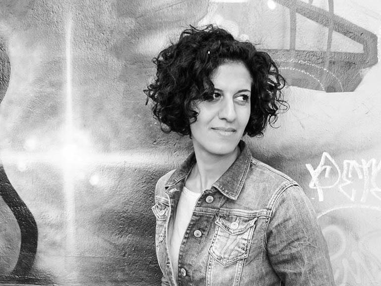 Die Schwarzweiß-Fotografie zeigt eine Frau mit lockigen, dunklen Haaren, die vor einer mit Graffiti besprühten Wand steht. Es die Musikerin und Kulturmanagerin Bahar Roshanai.