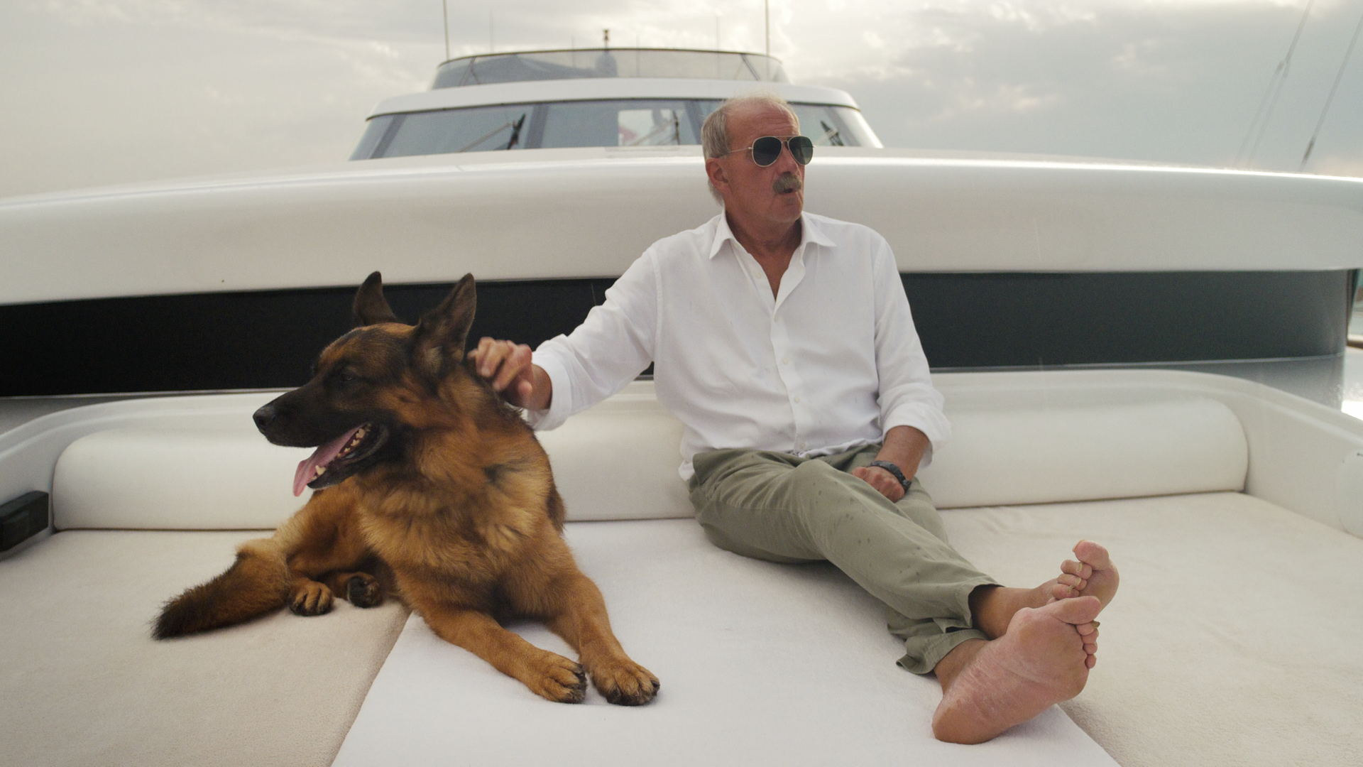 Szene aus der Netflix-Miniserie "Gunthers Millionen": Der Schäferhund Gunther sitzt auf einem weißen Polster an Deck einer Yacht, neben ihm ein Mann in heller Kleidung und mit Sonnenbrille. 