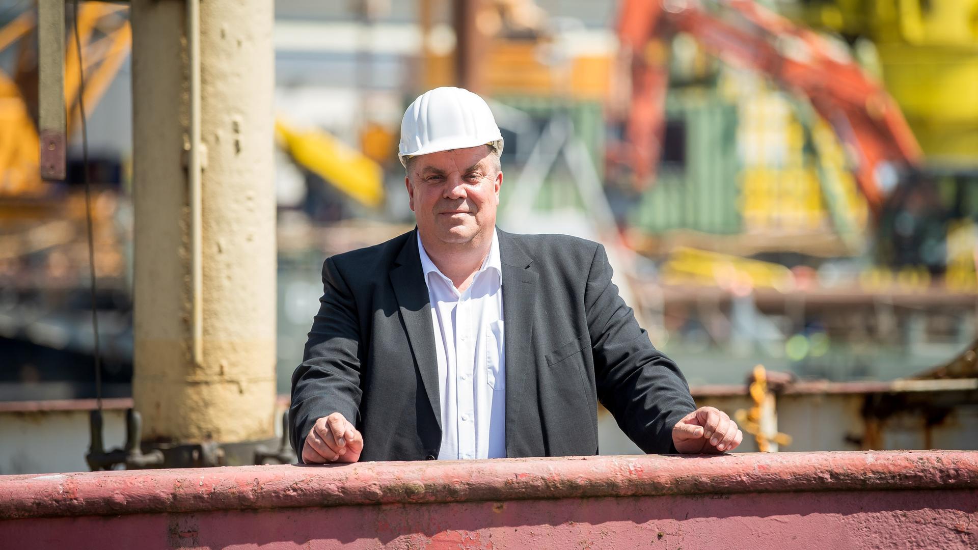 Carsten Feist, Oberbürgermeister der Stadt Wilhelmshaven steht mit Schutzhelm vor industrieller Hafenkulisse.