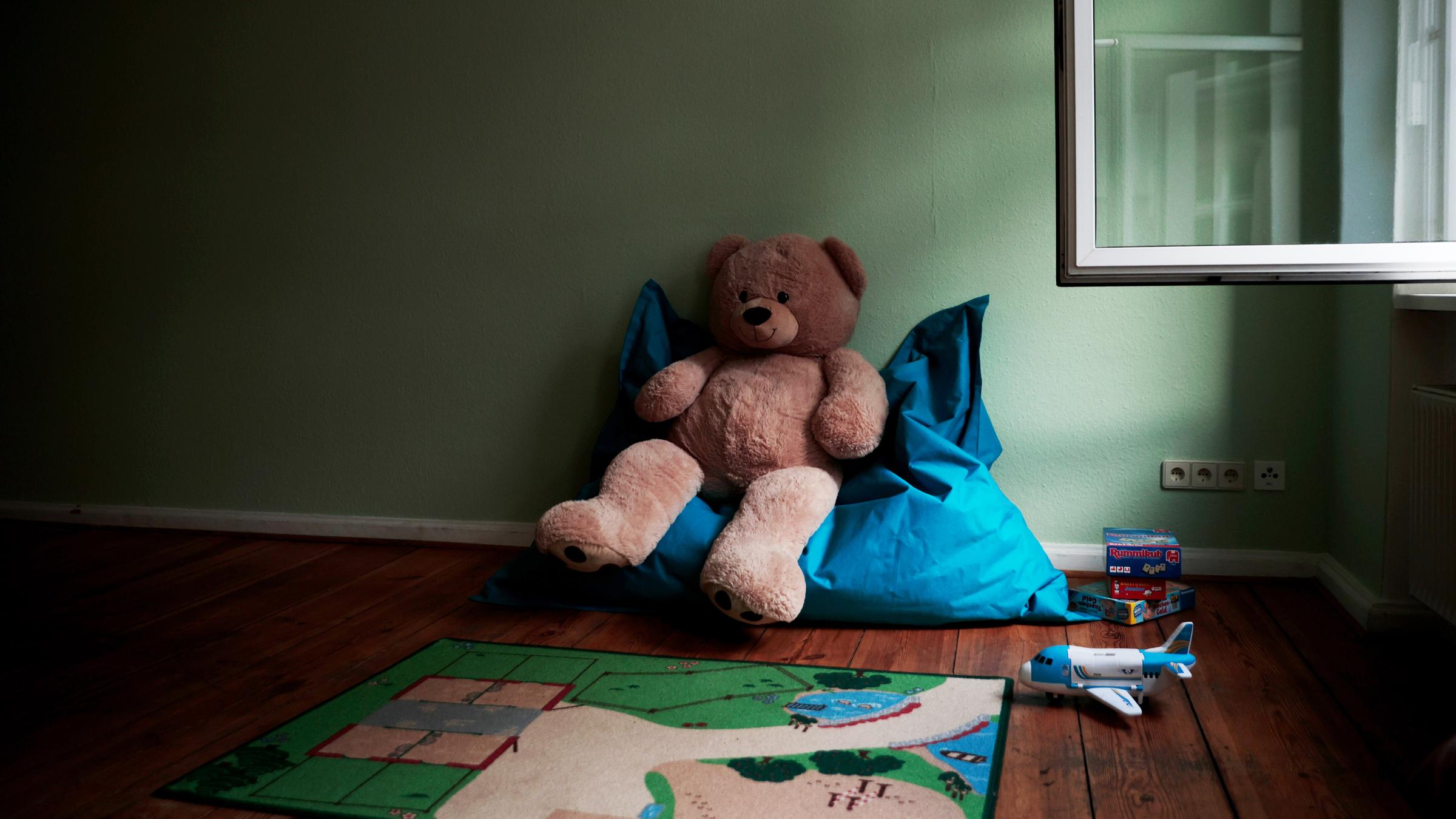 Ein großer Teddy sitzt auf einem Sitzsack in einem Spielzimmer. Daneben liegen weitere Spielsachen.