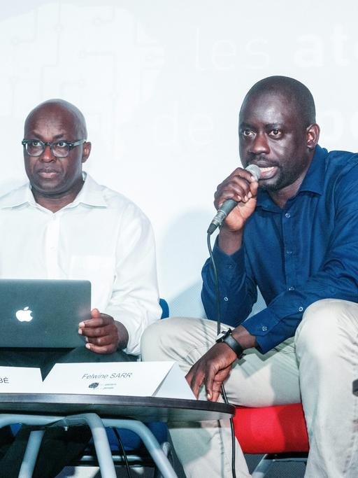 Achille Mbembe und Felwine Sarr sitzen an einem kleinen Tisch. Mbembe hat einen Laptop auf dem Schoß, Sarr spricht in ein Mikrofon.