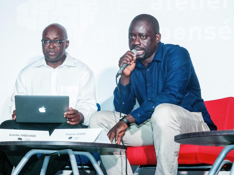 Achille Mbembe und Felwine Sarr sitzen an einem kleinen Tisch. Mbembe hat einen Laptop auf dem Schoß, Sarr spricht in ein Mikrofon.