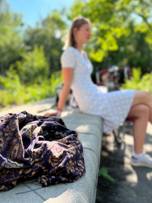 Die Studentin Nele sitzt in einem hellblauen Kleid auf einer Steinbank. Im Vordergrund liegt ihre Lieblingsdecke.