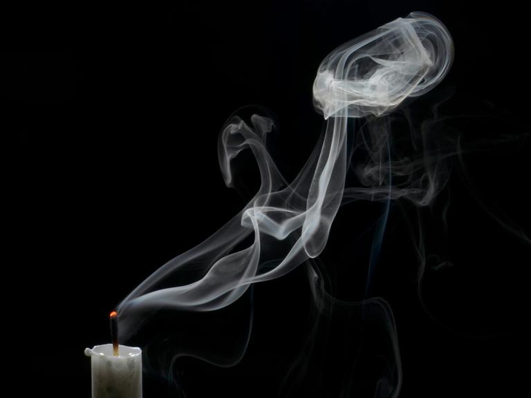 Eine soeben erloschene weiße Kerze vor scharzem Hintergrund. Der Docht glimmt noch und in der Mitte des Bildes steht weißer Rauch in der Luft.