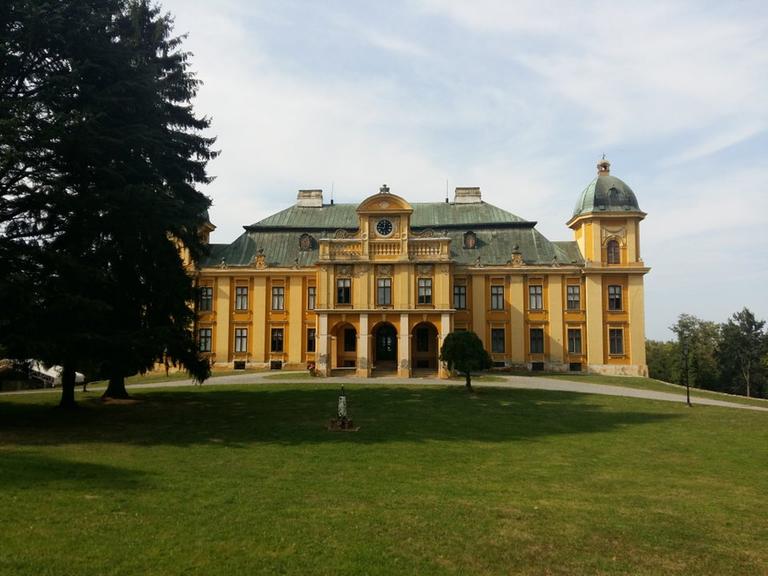 Blick auf ein vom Barock geprägten Schloss mit gelben Putz und imposanten Eingangsbereich, gekrönt von einer großen Uhr. Blick auf ein vom Barock geprägten Schloss mit gelben Putz und imposanten Eingangsbereich, gekrönt von einer großen Uhr.
