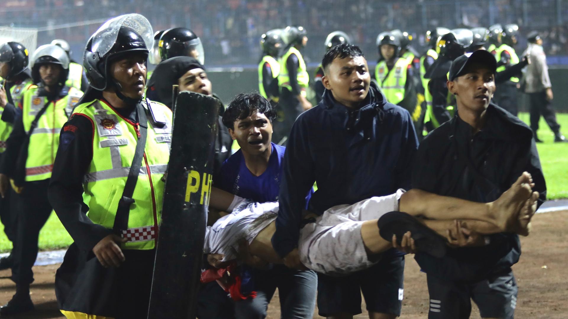 Fans tragen das Opfer am Spielfeldrand, daneben steht ein Polizist in gelber Warnweste mit einem Schild. Im Hintergrund auf dem rasen viele weitere Polizisten und eine Tribüne. 