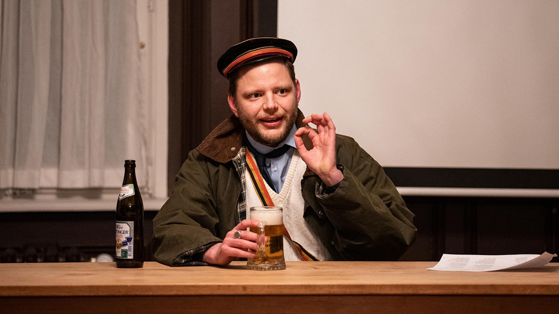 Im Bild aus der Inszenierung "saufen fechten heidelberg" sitzt ein Mann mit Uniformmütze an einem Tisch, hält ein Bierglas in der einen Hand und formt mit der anderen einen Ring aus Daumen und Zeigefinger.