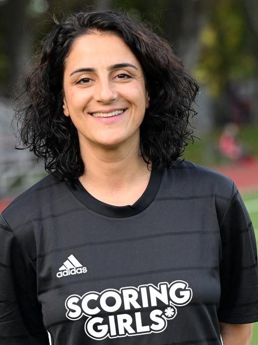Die Fußballerin Tugba Tekkal, Ex-Profi der Frauen-Bundesliga, steht auf einem Fußballplatz. Sie hat lockige, dunkle Haare trägt ein schwarzes Fußballshirt mit der Aufschrift "Scoring Girls".