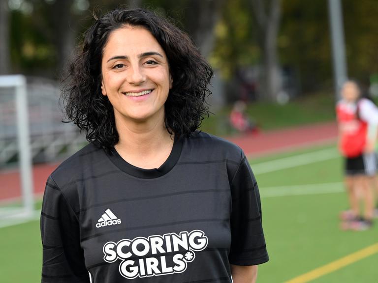 Die Fußballerin Tugba Tekkal, Ex-Profi der Frauen-Bundesliga, steht auf einem Fußballplatz. Sie hat lockige, dunkle Haare trägt ein schwarzes Fußballshirt mit der Aufschrift "Scoring Girls".