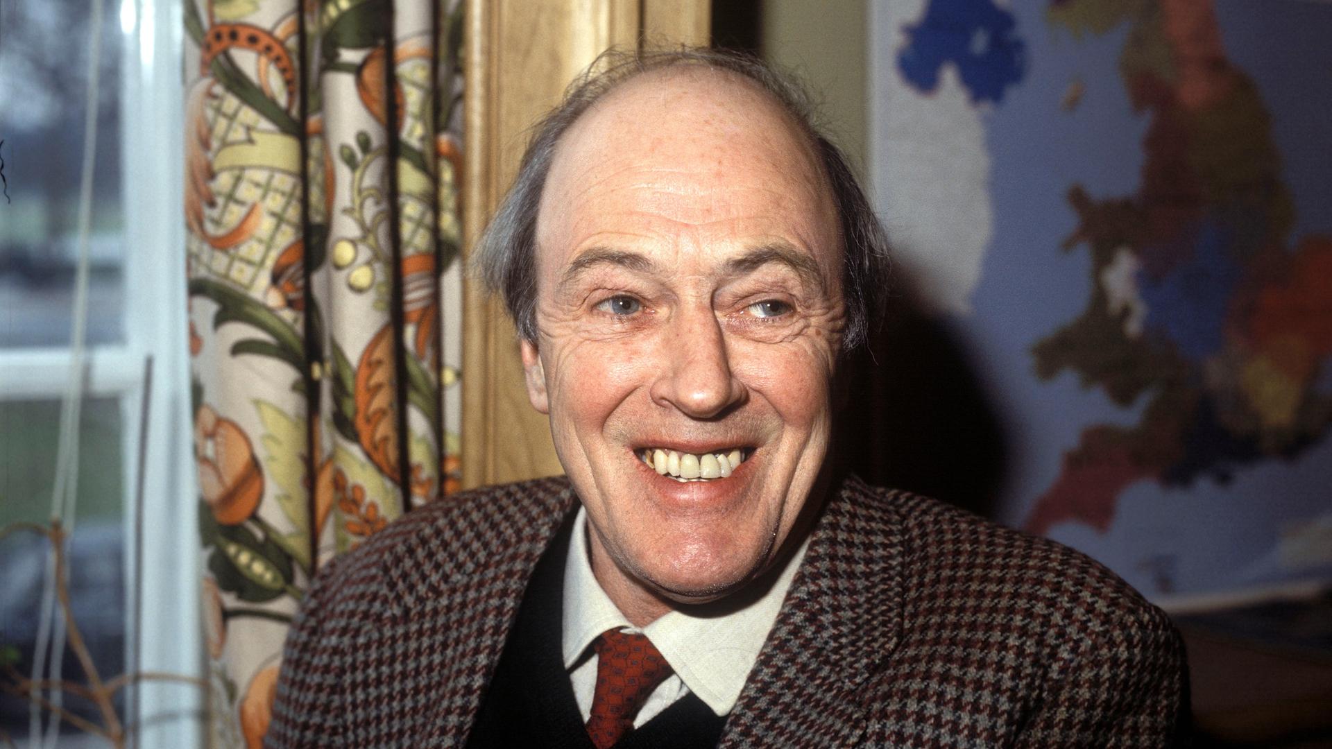 Roald Dahl trägt ein kariertes Jacket und lacht in die Kamera. Roald Dahl trägt ein kariertes Jacket und lacht in die Kamera.