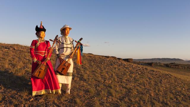 Zwei Frauen in mongolischer Nomadentracht stehen in der Weite der Wüste und halten zwei Pferdekopfgeigen in ihren Händen.