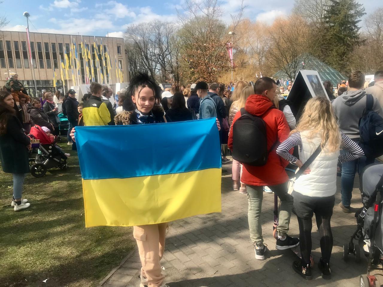 Eine junge Frau mit schwarzem Haar hält in einer Menschenmenge eine große ukrainische Flagge mit beiden Händen empor.