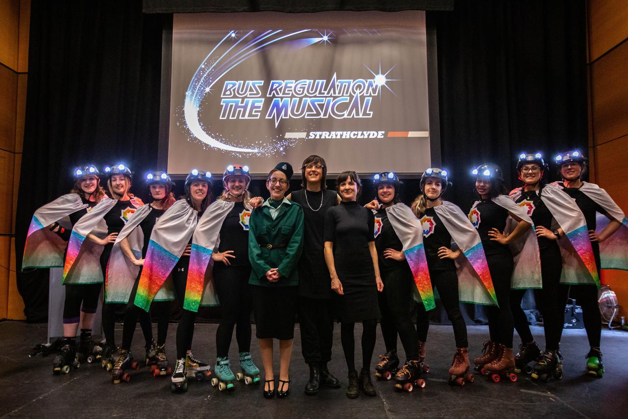 Auf der Bühne stehen 10 Darstellerinnen auf Rollschuhen in ihren Kostümen für das Musical "Bus-Regulation". Der Titel ist groß im Hintergrund zu sehen im Design von Starlight Express. In der Mitte ist Erfinderin Ellie Harrison. Auf der Bühne stehen 10 Darstellerinnen auf Rollschuhen in ihren Kostümen für das Musical "Bus-Regulation". Der Titel ist groß im Hintergrund zu sehen im Design von Starlight Express. In der Mitte ist Erfinderin Ellie Harrison.