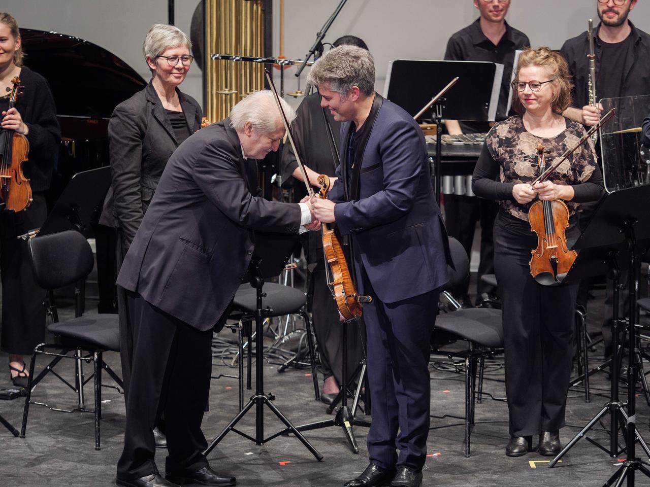 Auf einer Konzertbühne mit einem Ensemble im Hintergrund bedankt sich ein älterer Herr mit einer Verbeugung und Händedruck bei einem Mann mit einer Geige in der Hand. 