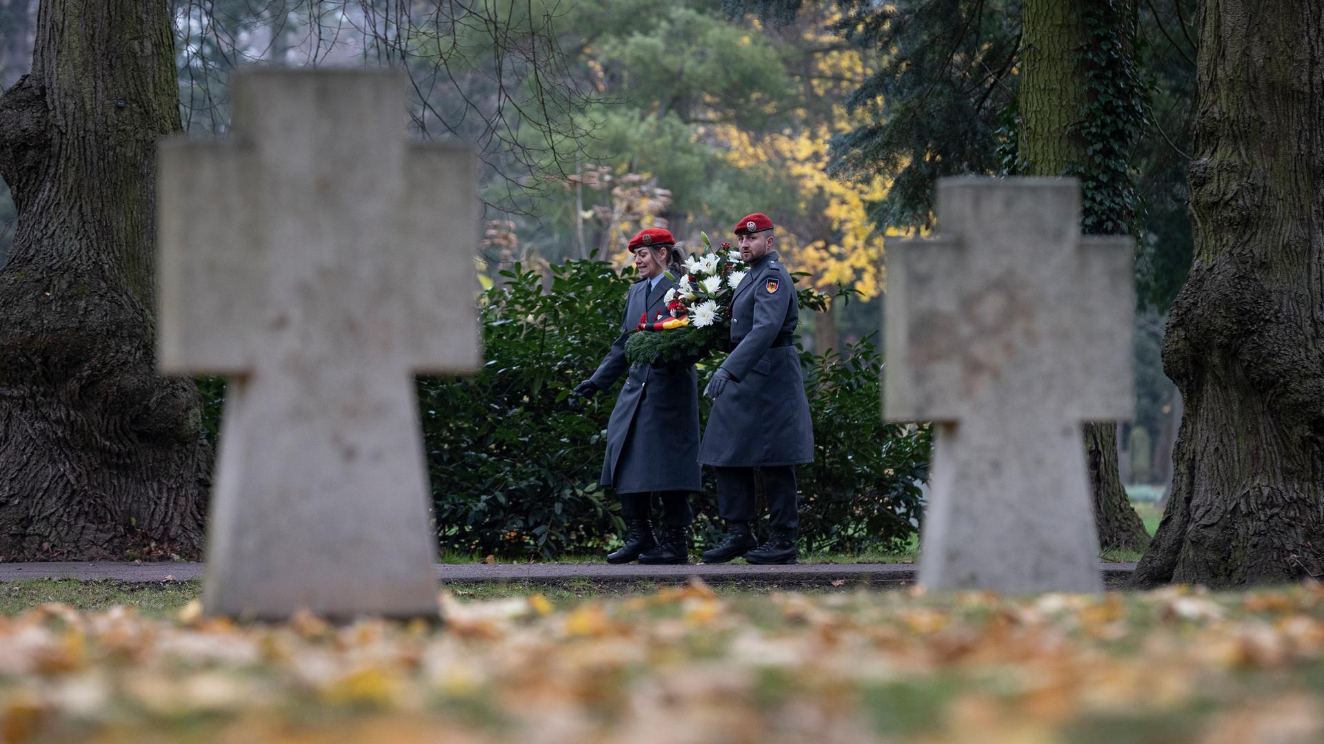 Ein Friedhof im Herbst: Im Fokus sind ein Soldat und eine Soldatin in Uniform, die zum Volkstrauertag einen Kranz tragen. Im Vordergrund unscharf zwei Grabkreuze und Rasen mit Laub.