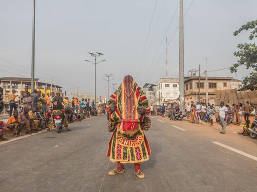 Ein Egoun steht auf einer Straße in Porto Novo: Ein Mann, in bunte weite Stoffe gewandet, die auch das Gesicht verhüllen.