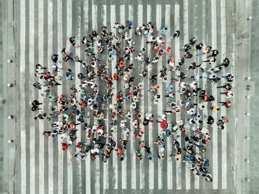 Vogelperspektive auf eine Gruppe von Menschen, die auf einem Zebrastreifen steht und als Masse die Form einer Sprechblase abbilden. Wird da Dialekt gesprochen?
