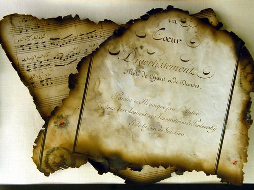 Zwei Blätter, die rundherum Spuren von Feuer zeigen. Es ist ein Deckblatt, auf dem unter anderem "Divertissement" steht und dass dieses Heft Noten und Verse enthält. Dahinter ist das zweite Blatt zu sehen, es zeigt ein paar handschriftliche Noten.