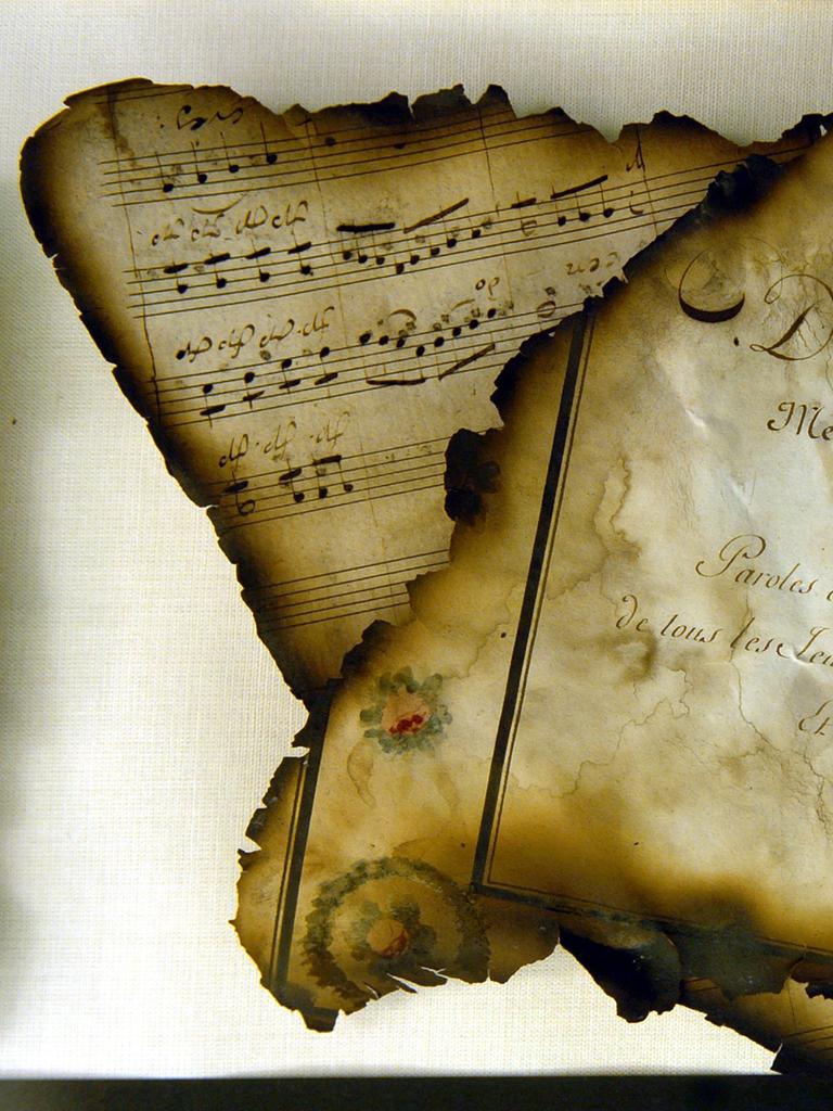 Zwei Blätter, die rundherum Spuren von Feuer zeigen. Es ist ein Deckblatt, auf dem unter anderem "Divertissement" steht und dass dieses Heft Noten und Verse enthält. Dahinter ist das zweite Blatt zu sehen, es zeigt ein paar handschriftliche Noten.