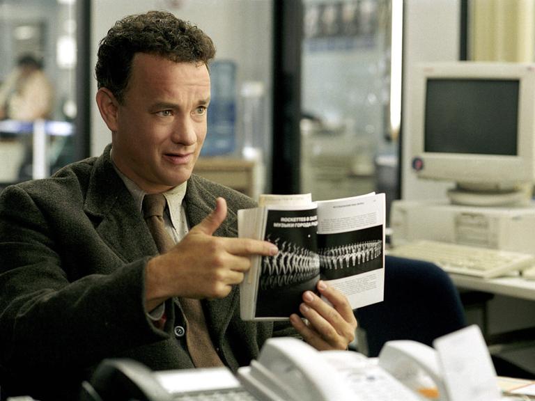 Der Schauspieler Tom Hanks zeigt im Film "Terminal" mit dem Finger auf die Seite eines Buches, die ein Foto einer Reihe von Tänzerinnen zeigt. Im Hintergrund idt ein Computer zu sehen.