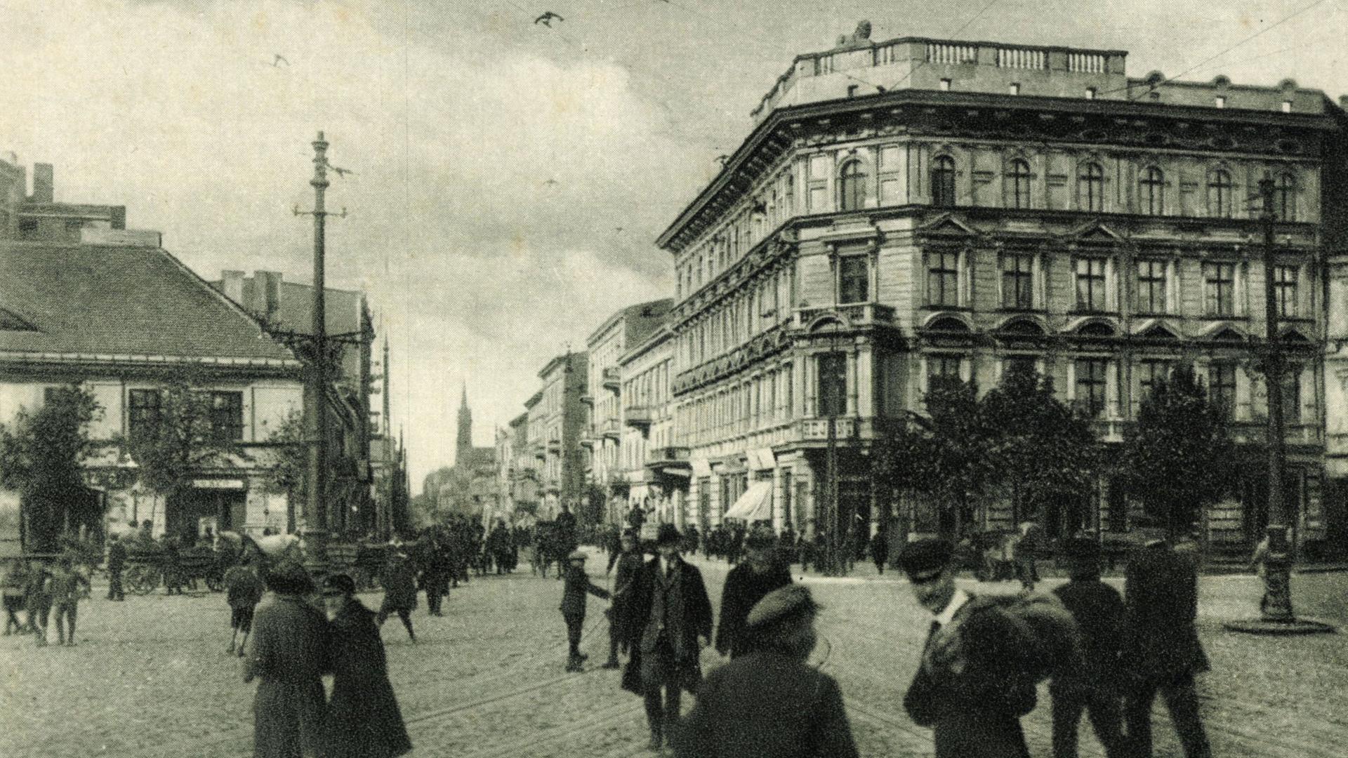 Schwarz-Weiß-Bild eines Gebäudes am Freiheitsplatz in Lodz. Auf dem Platz sind mehrere Menschen zu sehen, die den Platz kreuzen. 