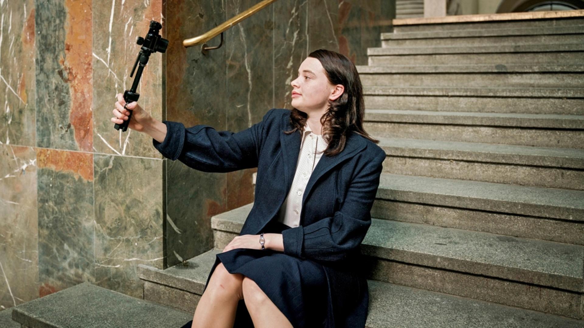 Eine junge Frau sitzt auf einer Treppe und filmt sich mit einer Kamera.