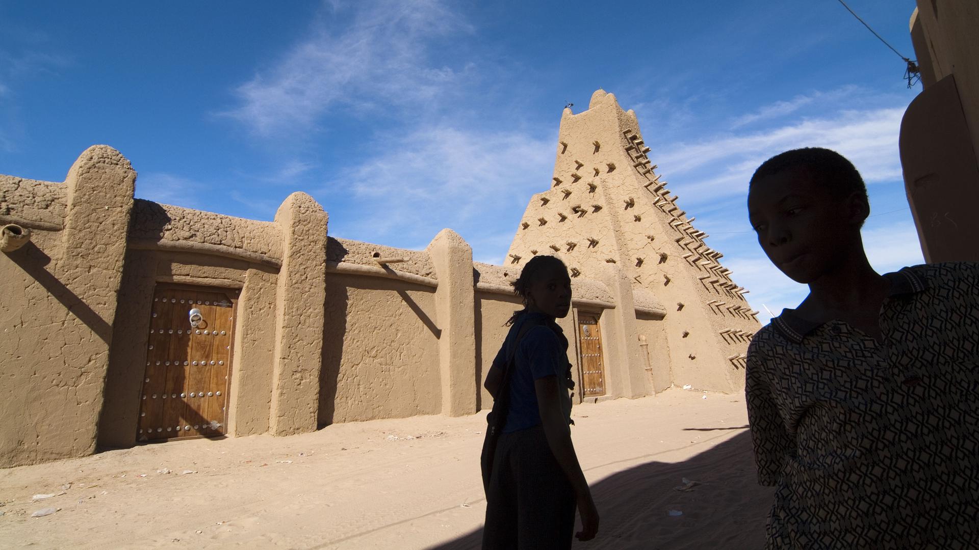 Blick auf die in Lehm gebaute Moschee vor blauem Himmel. Davor die Silhoutte zweier Kinder.