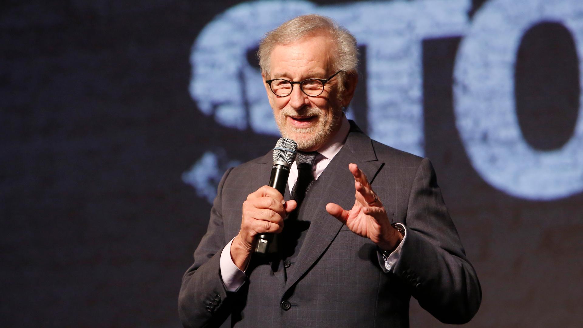Steven Spielberg hält auf einer Bühne ein Mikrofon in der Hand. Im Hintergrund ist ein Teil des Schriftzugs "West Side Story" zu erkenenn. Steven Spielberg hält auf einer Bühne ein Mikrofon in der Hand. Im Hintergrund ist ein Teil des Schriftzugs "West Side Story" zu erkenenn.