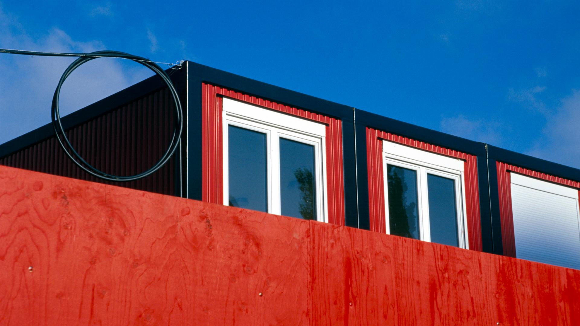 Hinter einer roten Wand ist der obere Teil eines Wohncontainers zu erkennen.